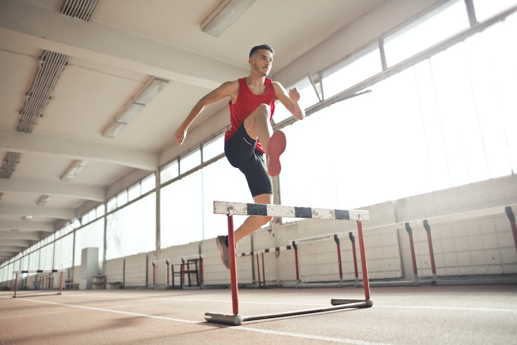 Powerful male athlete jumping over hurdle on running track while training at sports hall